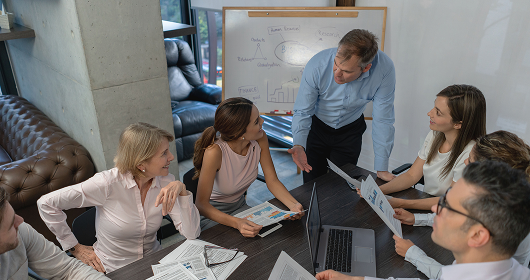 Manager leading a team meeting around a table with charts on a whiteboard
